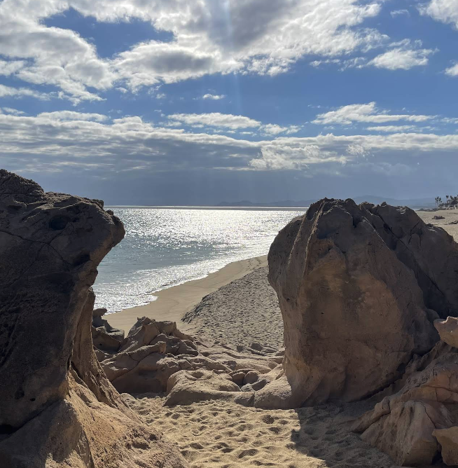 Two weathered rock formations frame a sandy path to a sunlit sea; the calm beach extends to the horizon as sparkling water reflects sunlight under a blue, partly cloudy sky.