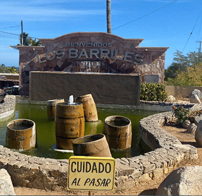 Object: circular stone fountain with wooden barrels arranged in a ring. Action: water pools around barrels. Context: backdrop features a stone wall sign reading BIENVENIDOS LOS BARRILES; a yellow sign reads CUIDADO AL PASAR; blue sky. Transcriptions: BIENVENIDOS LOS BARRILES CUIDADO AL PASAR