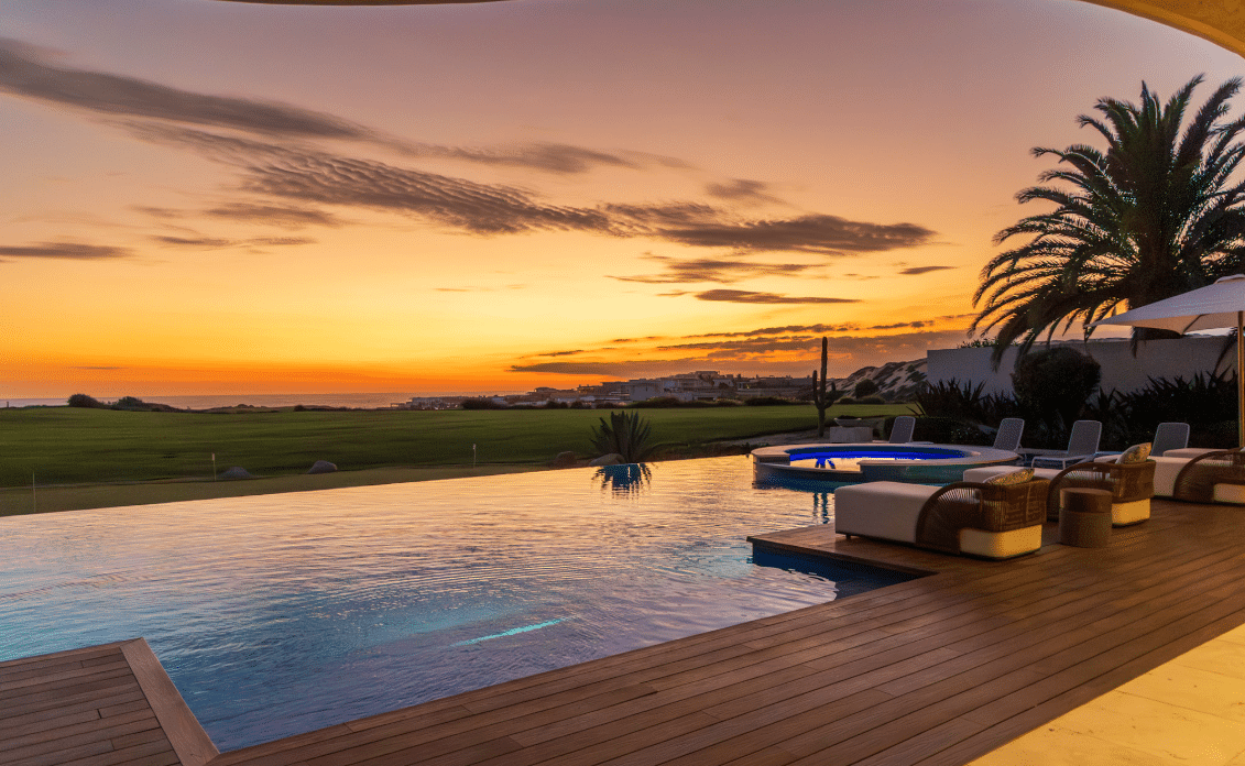 An infinity pool on a deck reflects a warm sunset. Lounge chairs with wicker seating line the deck, palm trees nearby, and a distant grassy landscape stretches to the horizon.