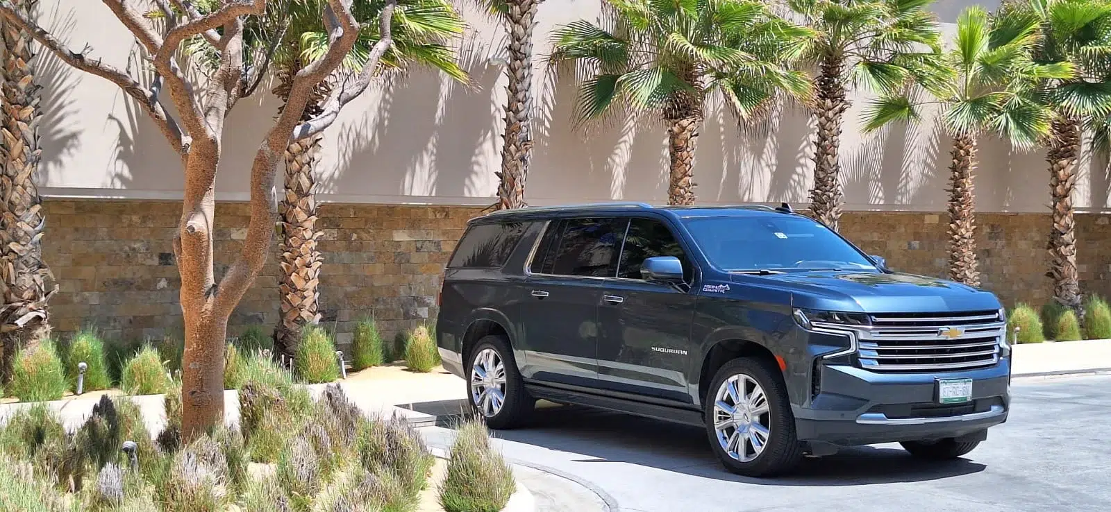 all inclusive airport shuttle A dark gray Chevrolet Suburban is parked along a curved driveway; the side badge reads SUBURBAN. Setting features a stone wall, palm trees, and drought-tolerant landscaping.